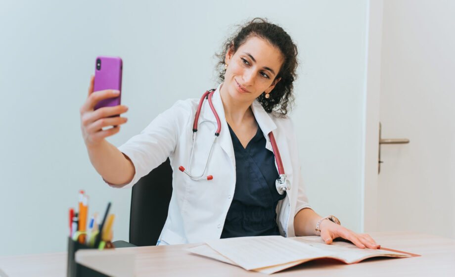 L'immagine rappresenta la foto di una donna medico mentre tiene in mano un cellulare rosa, per gestire le attività di social media