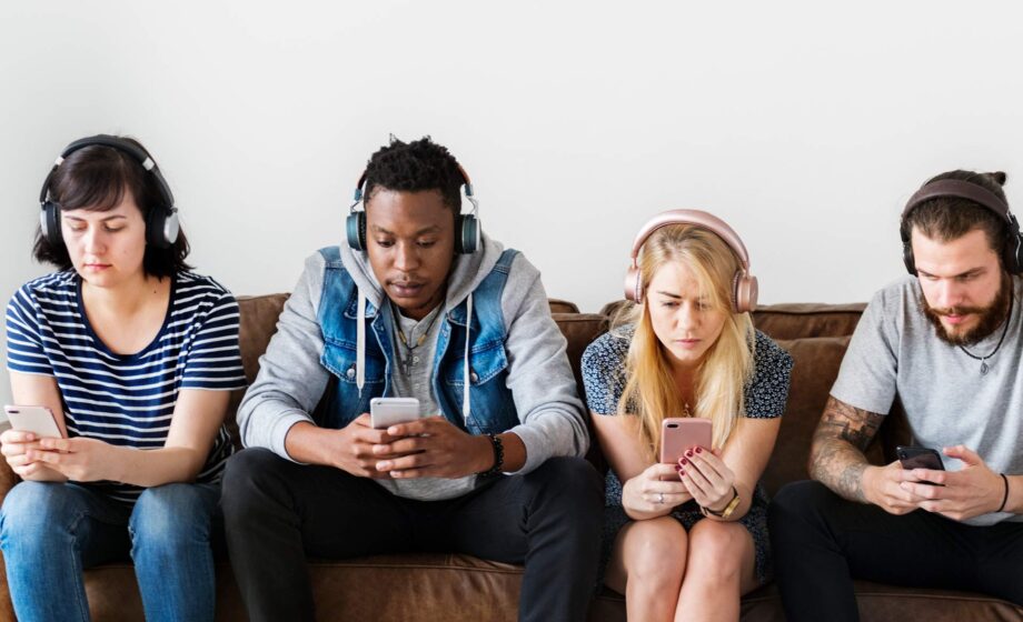 Quattro ragazzi seduti su un divano mentre guardano ossessivamente il cellulare che tengono in mano, affetti da dipendenza digitale e comportamentale